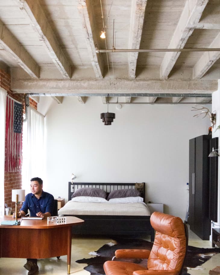 Loft bedroom with exposed beams, leather chairs, a wooden desk, and a bed with gray bedding. A person sits at the desk.