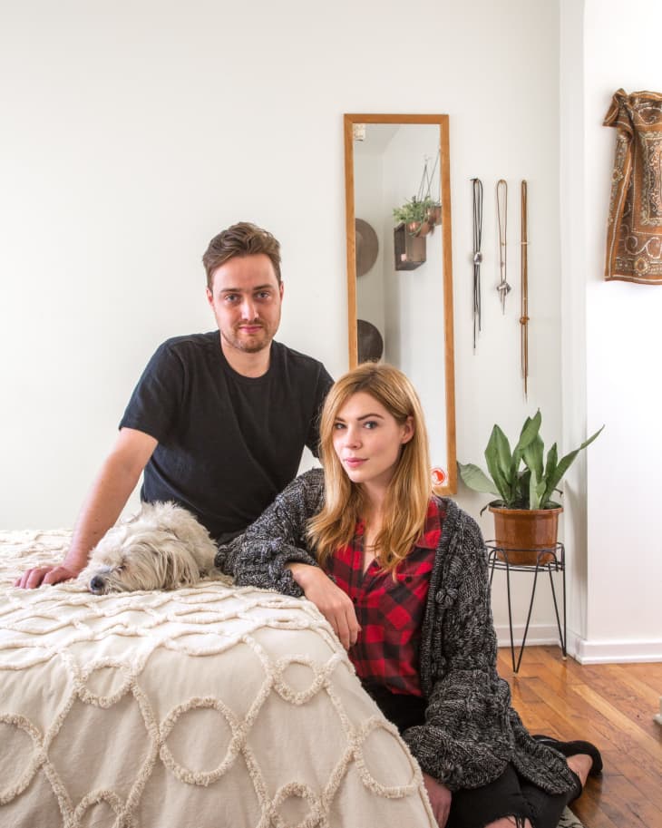 Man and woman sitting on a bed with a dog, next to a mirror and potted plant in a cozy room.