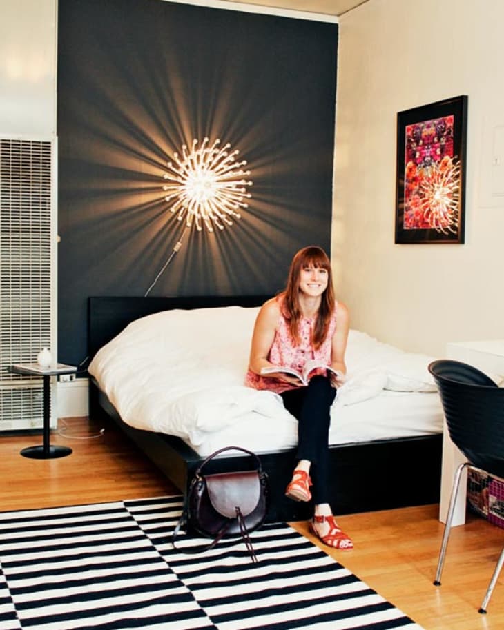 Woman sitting on bed with book, modern light fixture, striped rug, and wall art in cozy bedroom.