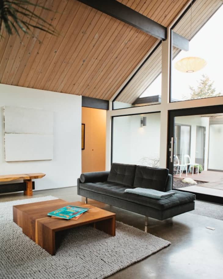Modern living room with vaulted wood ceiling, black sofa, wooden coffee table, and large windows overlooking a patio.