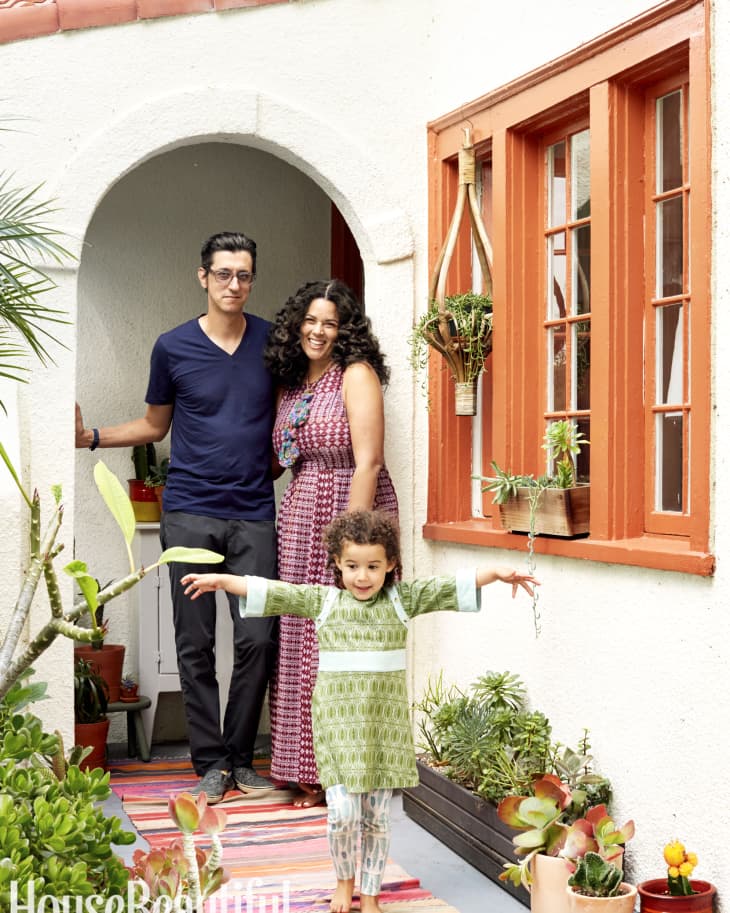 Family standing in a colorful patio with potted plants and a child in a green dress, smiling under an archway.
