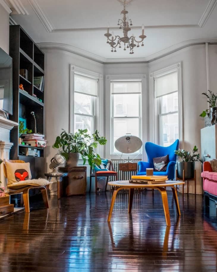 Living room with a blue armchair, wooden coffee table, plants, and a chandelier, featuring large bay windows.