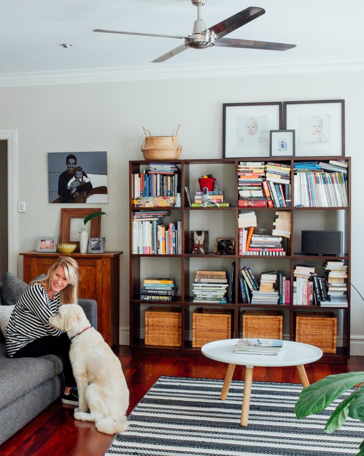 Living room with a woman petting a dog on a gray sofa, bookshelves, and a round coffee table on a striped rug.