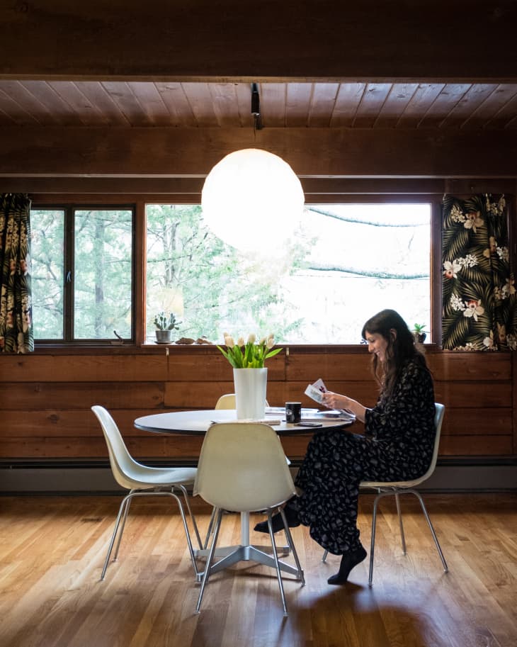 Woman reading at a round table with white chairs, wooden walls, and floral curtains, under a large spherical light.