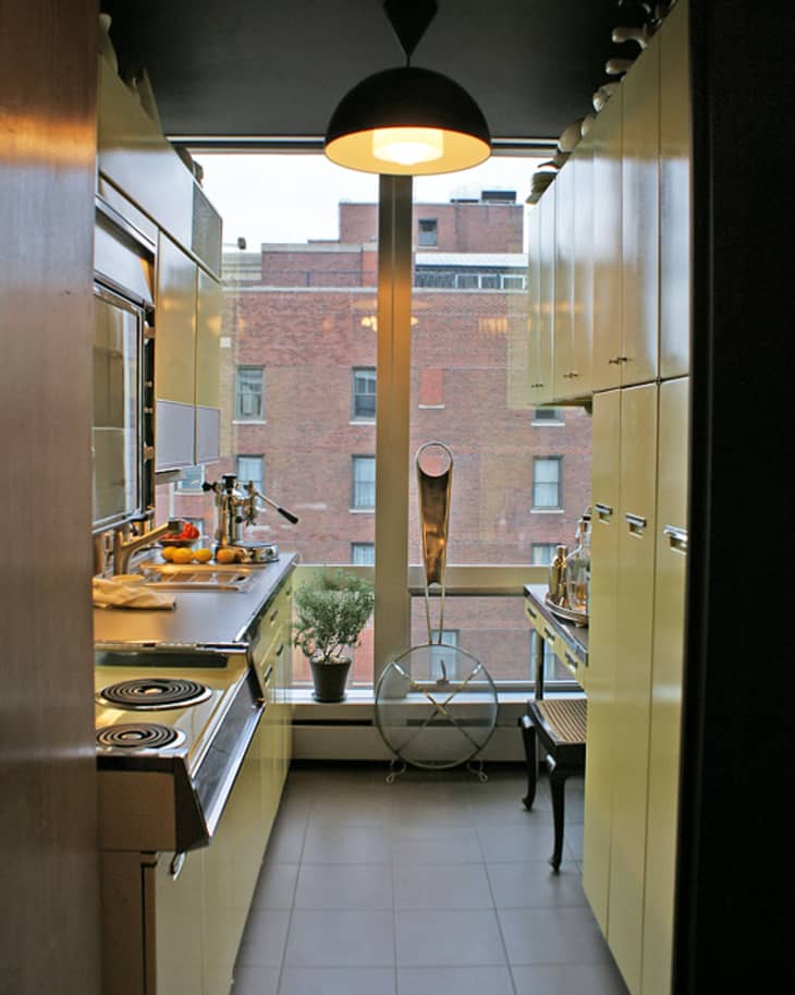 Narrow kitchen with yellow cabinets, stainless steel appliances, and a large window overlooking brick buildings.