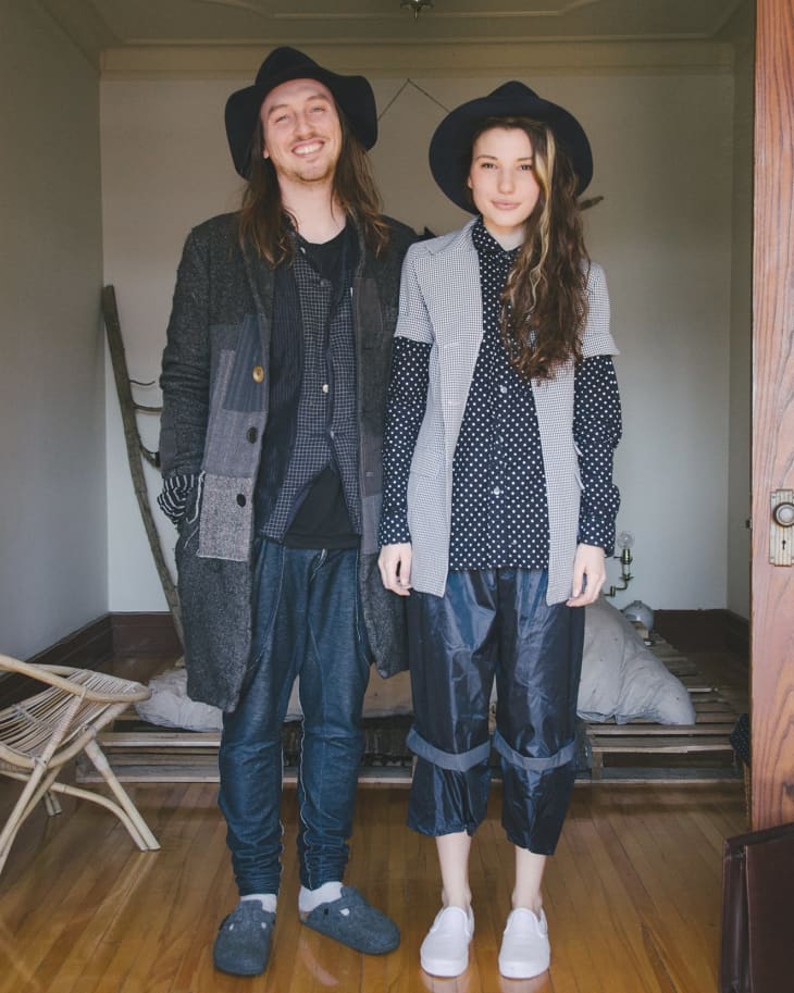Two people in casual, layered outfits and hats standing on a wooden floor in a minimalist room.