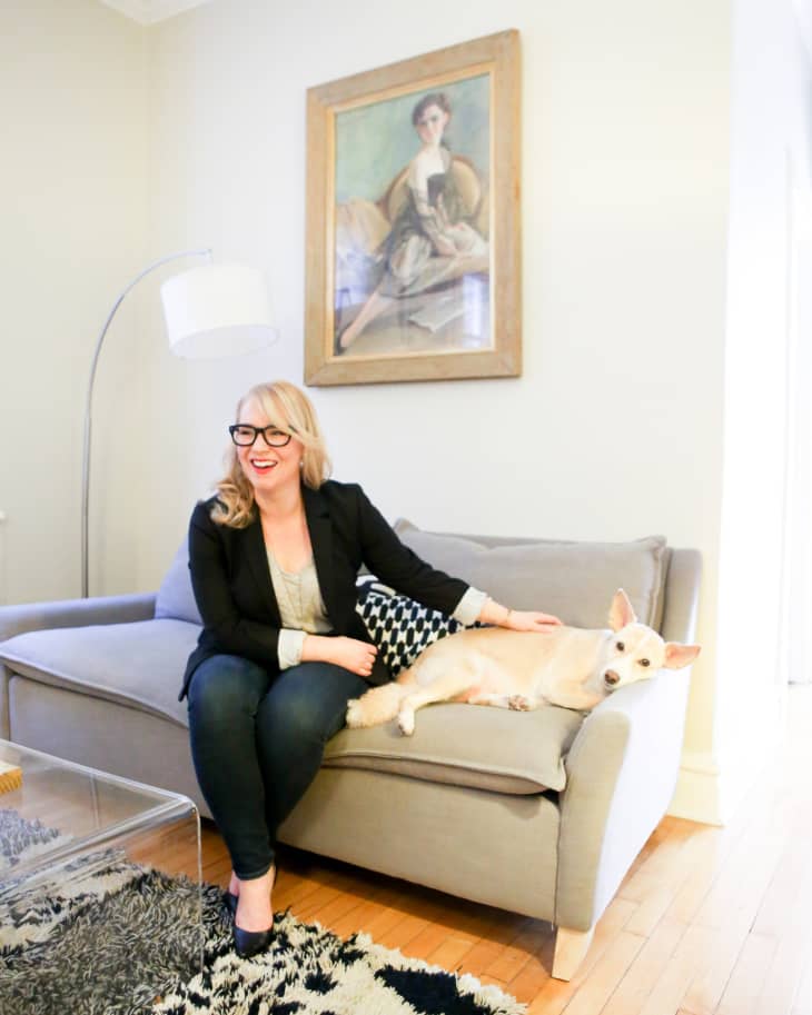 Woman sitting on a gray sofa with a small dog, next to a modern floor lamp and abstract painting.