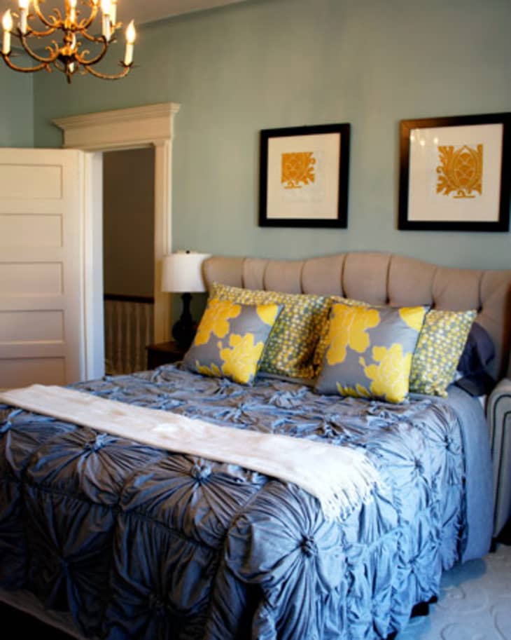 Bedroom with a tufted headboard, blue bedding, yellow floral pillows, chandelier, and framed art on light blue walls.
