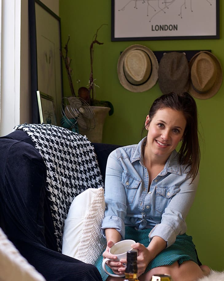 Woman in denim shirt holding a mug, sitting on a sofa with houndstooth blanket, green wall with hats and London map.
