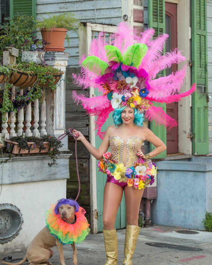Woman in colorful feathered costume with matching dog on porch, both wearing vibrant headpieces and ruffled collars.