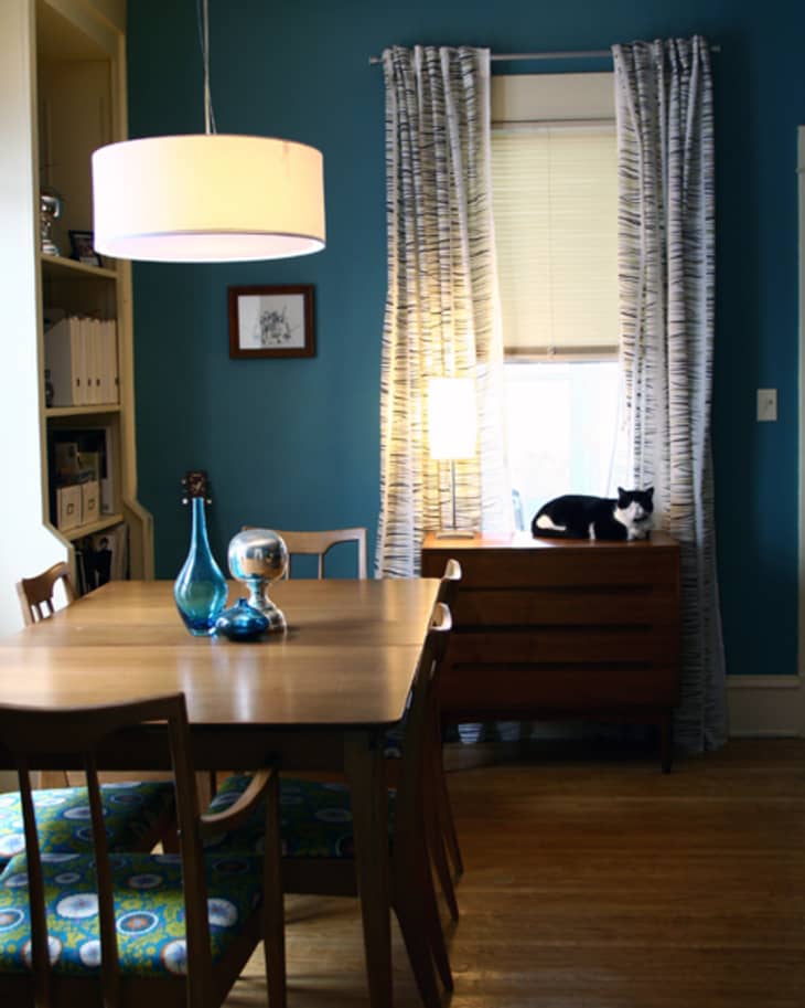 Dining room with wooden table, blue walls, patterned chairs, pendant light, and a cat on a dresser by the window.