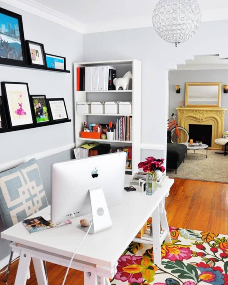 Home office with white desk, iMac, colorful floral rug, bookshelf, and view into living room with yellow fireplace.