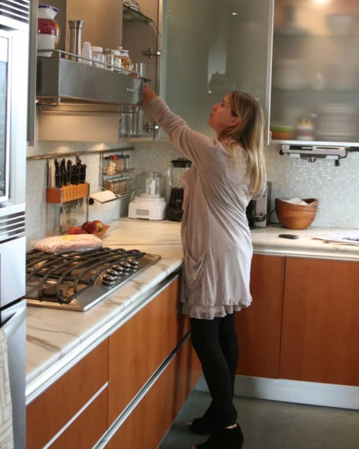 Woman reaching for items in a modern kitchen with wooden cabinets, marble countertops, and stainless steel appliances.