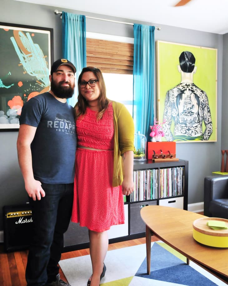 Couple standing in a colorful living room with vibrant artwork, blue curtains, and a record collection.