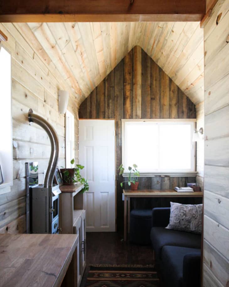 Cozy cabin interior with wood paneling, black sofa, desk, and potted plants by a window.