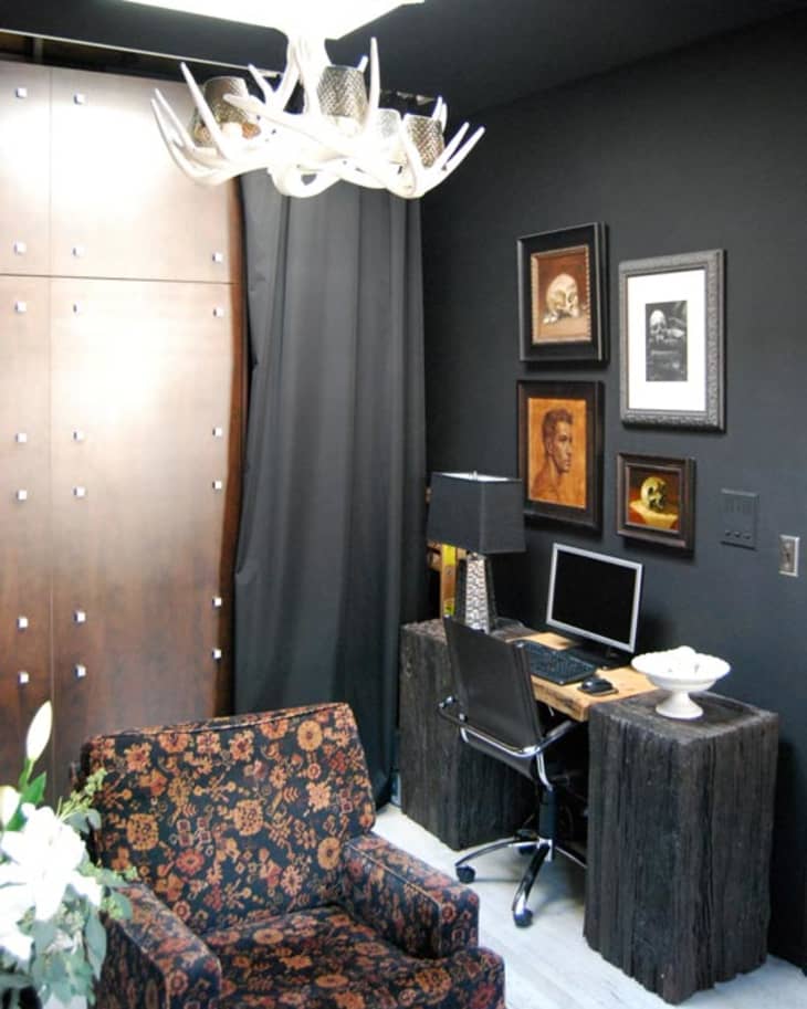 Home office with black walls, floral armchair, antler chandelier, framed art, and a computer desk with a black lamp.