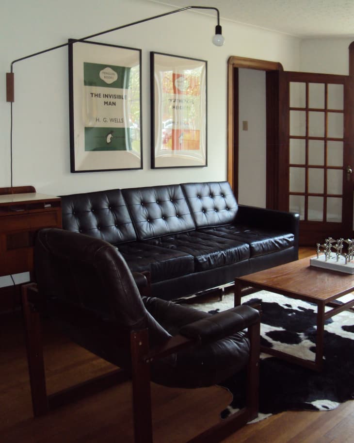 Mid-century modern living room with black leather sofa, armchair, wooden coffee table, and framed book cover art.