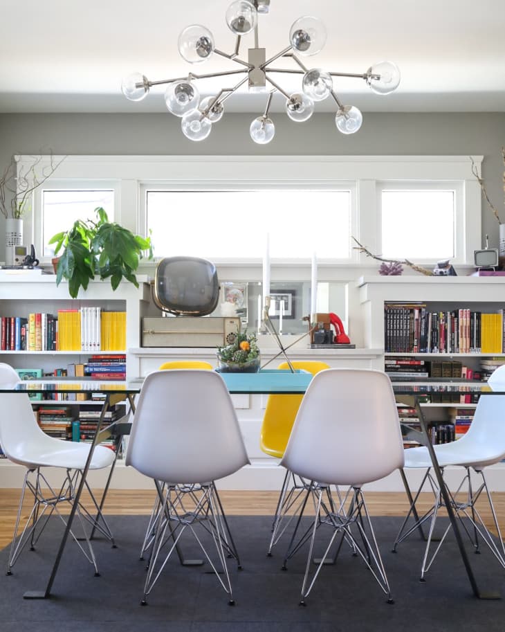 Dining room with modern chandelier, glass table, white and yellow chairs, bookshelves, and a vintage TV.