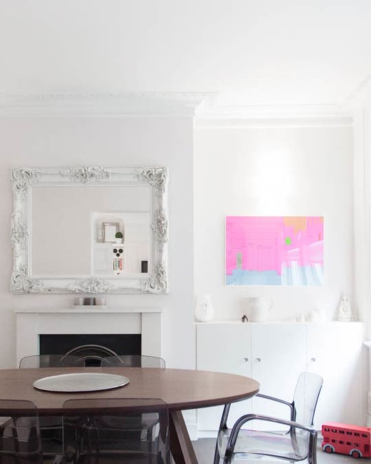 Dining room with oval wooden table, transparent chairs, ornate mirror, and bright pink abstract painting.