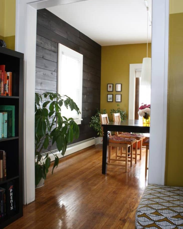Dining room with black accent wall, wooden floor, black table, wooden chairs, large plant, and bookshelf.