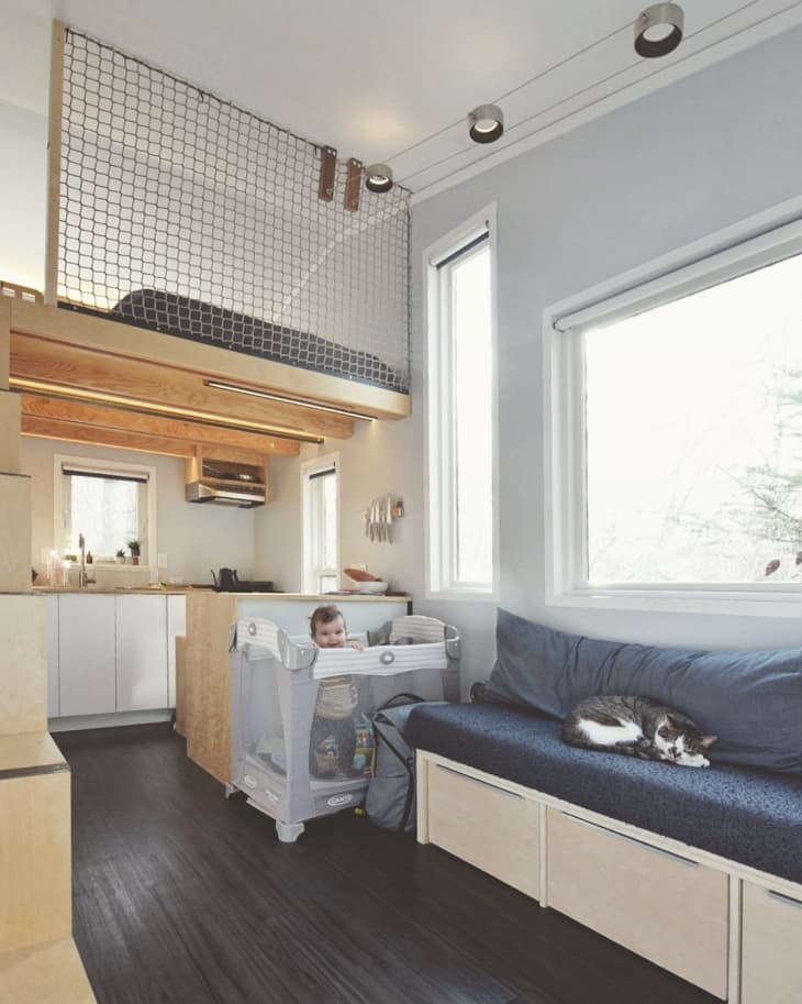 Baby in playpen and cat on blue sofa in modern tiny home with loft bed and kitchen area.