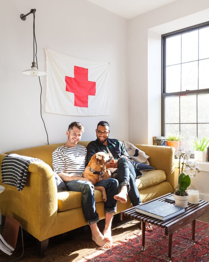 Two people sitting on a yellow sofa with a dog, under a Red Cross flag, next to a window and a wooden coffee table.