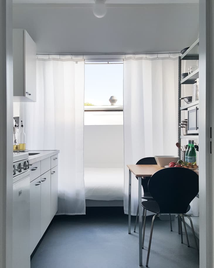 Small kitchen with white cabinets, a wooden table, black chairs, and a window with sheer curtains.