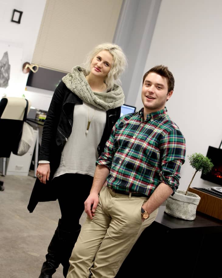 Two people standing in an office, one in a plaid shirt and the other in a scarf, with computers and decor in the background.