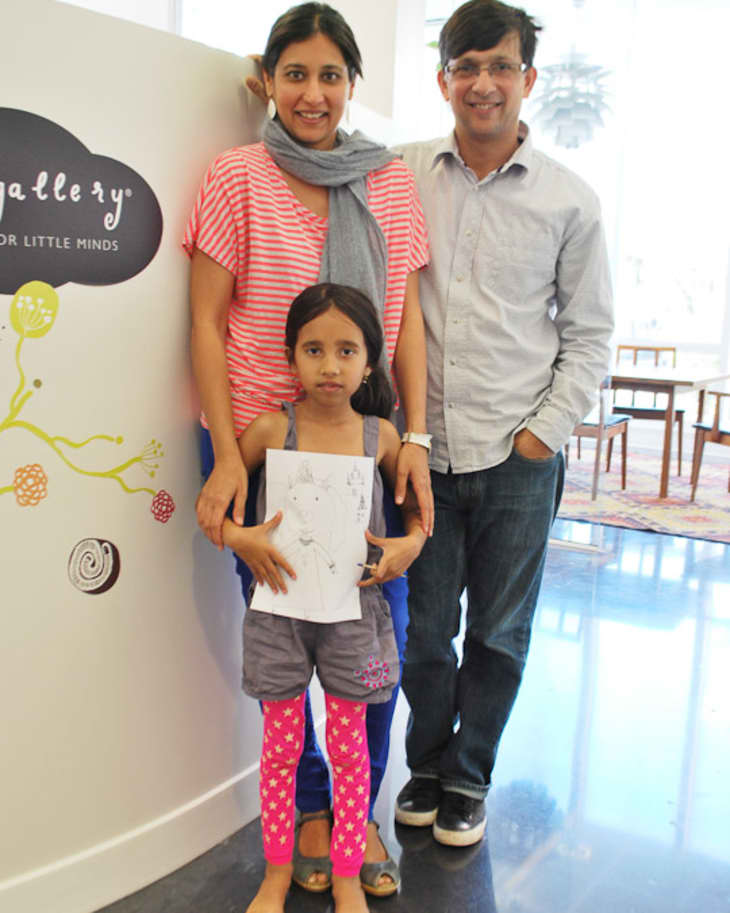 Family posing with a child's drawing in an art gallery, featuring a woman in a striped shirt, a man in a light shirt, and a girl in pink leggings.
