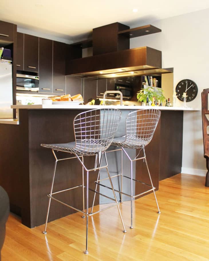 Modern kitchen with dark cabinets, wire bar stools, wooden floor, and a clock on the wall.