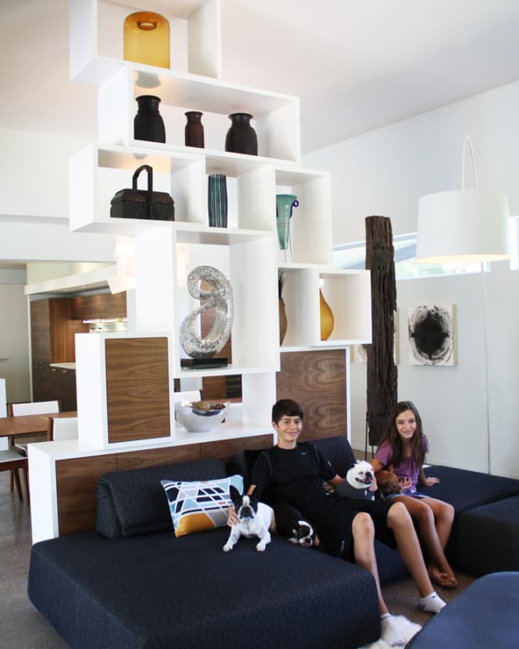 Teenagers sitting on a dark sofa with two dogs, surrounded by modern white shelving with decorative vases and sculptures.