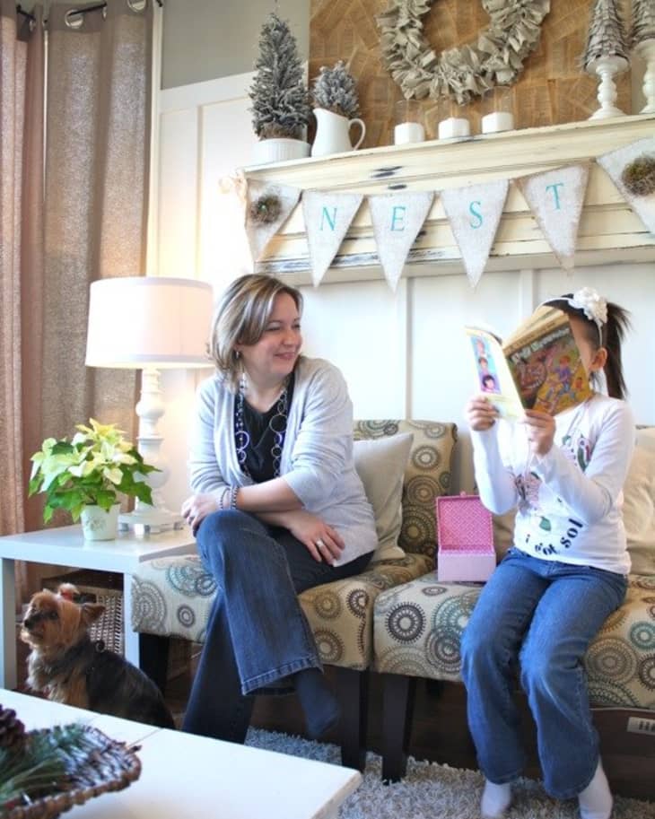 Woman and child sitting on a patterned sofa, child reading a book, with a dog nearby and festive decor on the mantel.