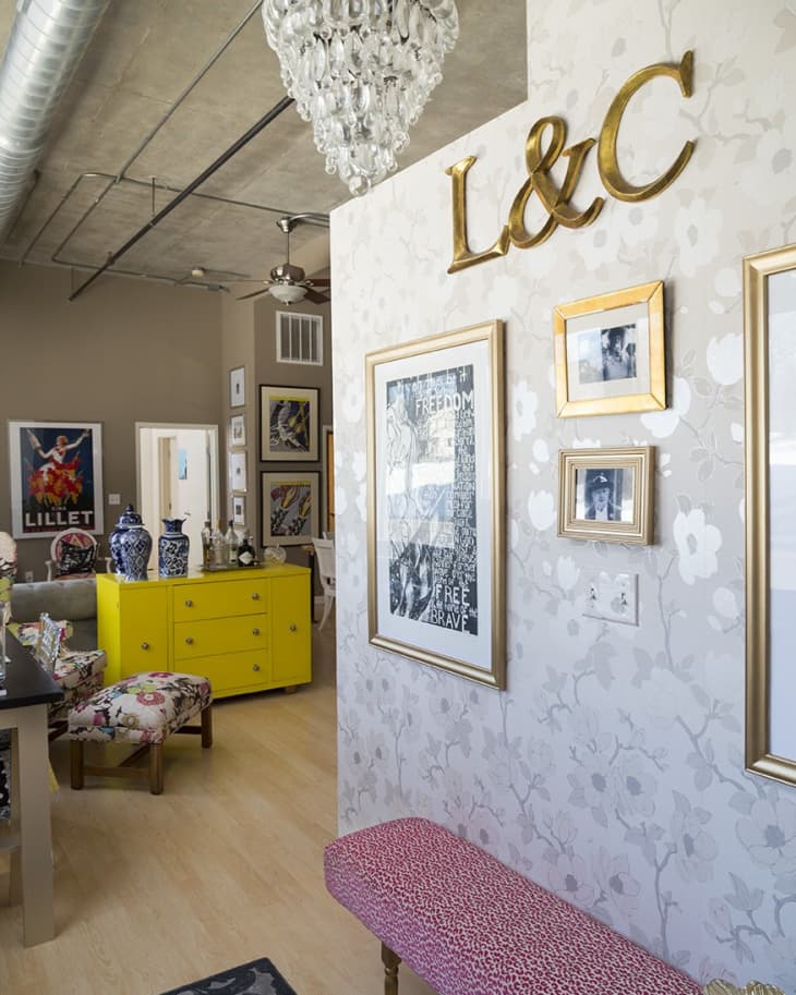 Chic living room with floral wallpaper, crystal chandelier, yellow dresser, and pink bench. Art and decor add vibrant touches.