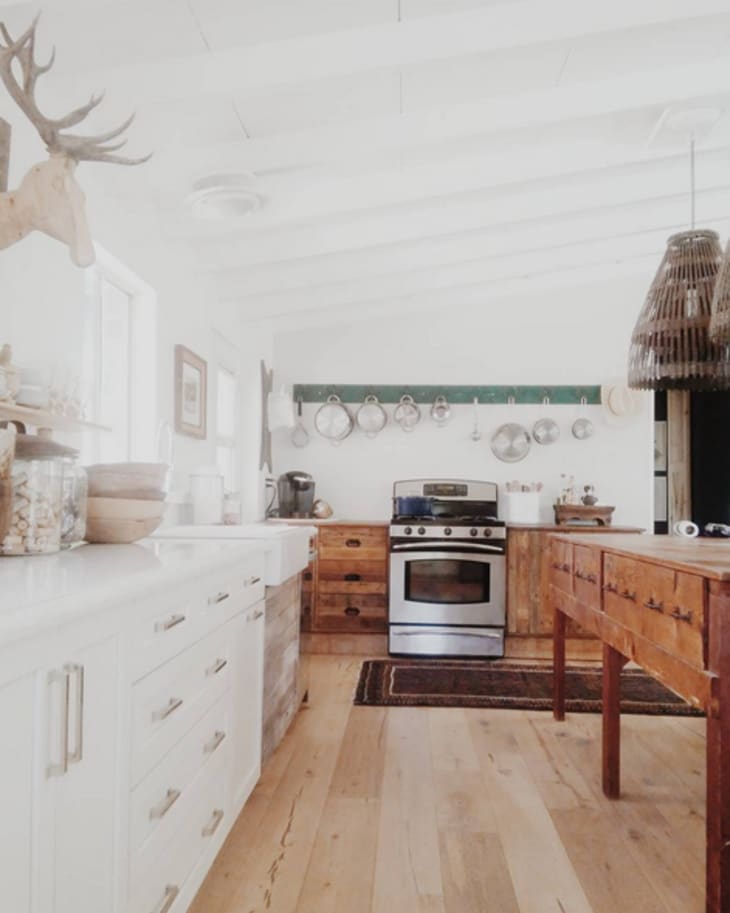 Rustic kitchen with wooden island, stainless steel stove, hanging pots, and white cabinets under a beamed ceiling.