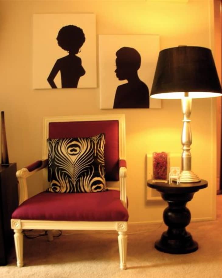 Red armchair with a patterned cushion beside a black lamp on a round table, under silhouette art on the wall.