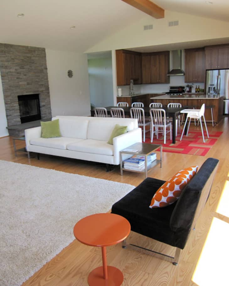Open-plan living room with white sofa, black chair, orange table, and kitchen with wooden cabinets and metal bar stools.
