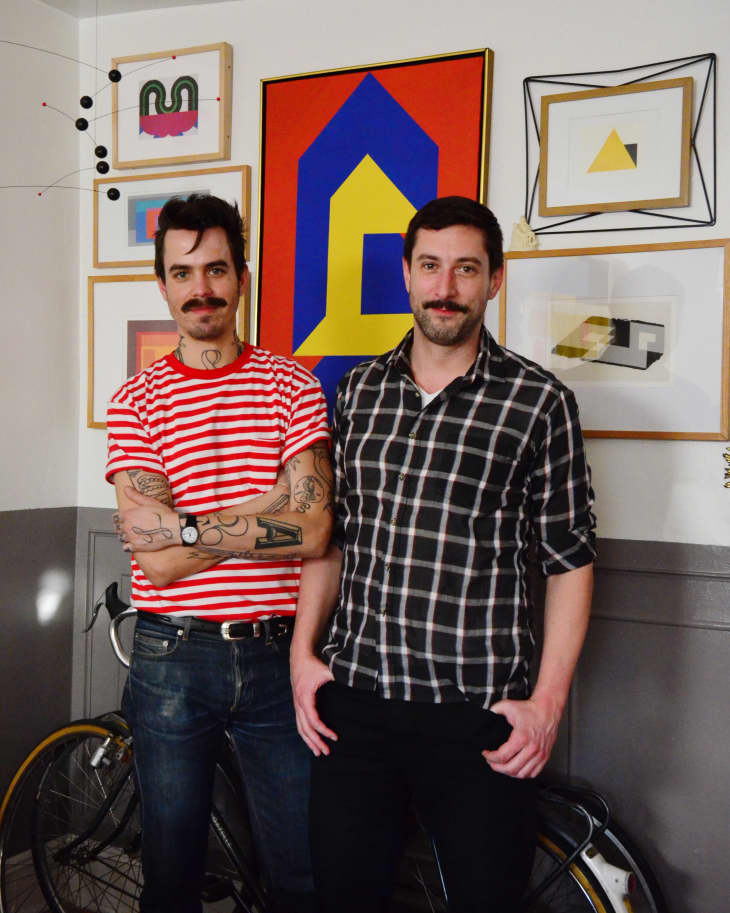 Two men standing in front of a wall with colorful geometric art, one in a red striped shirt, the other in a plaid shirt, with a bicycle.