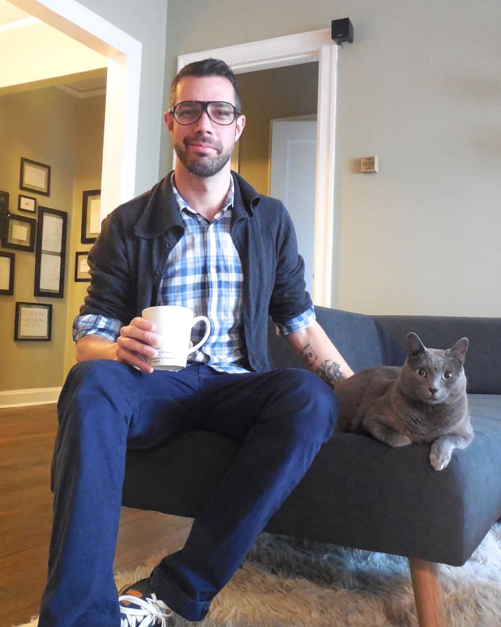 Man in glasses holding a mug, sitting on a gray sofa with a gray cat beside him in a cozy living room.