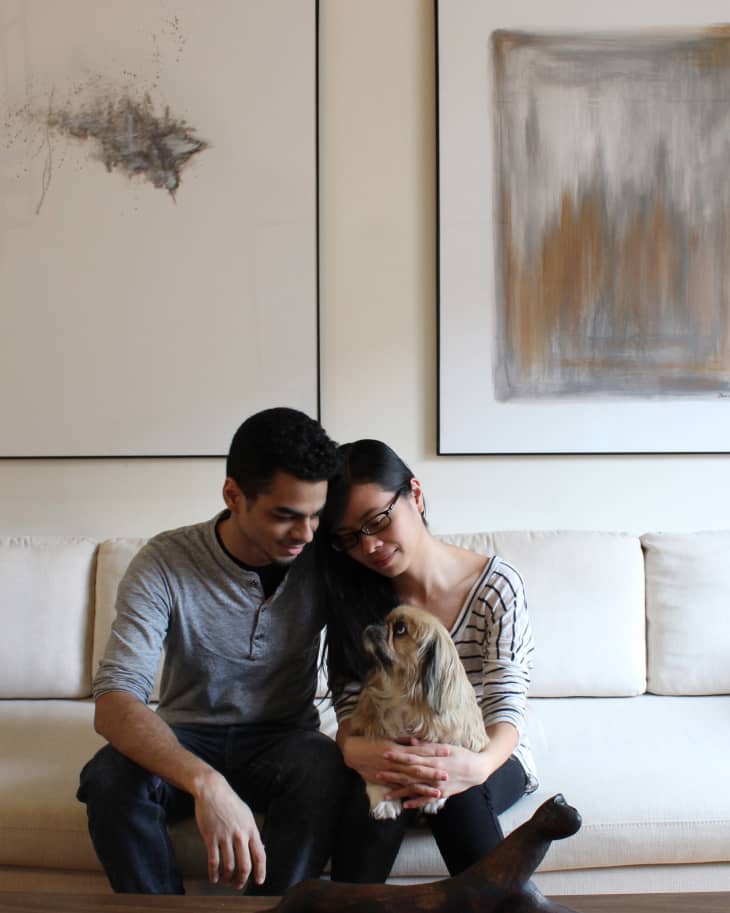 Couple sitting on a cream sofa with a small dog, surrounded by abstract wall art and a wooden sculpture on the table.