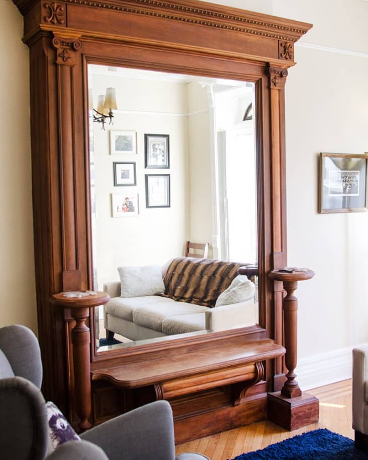 Large ornate wooden mirror reflecting a cozy living room with a beige sofa, brown throw, and framed wall art.