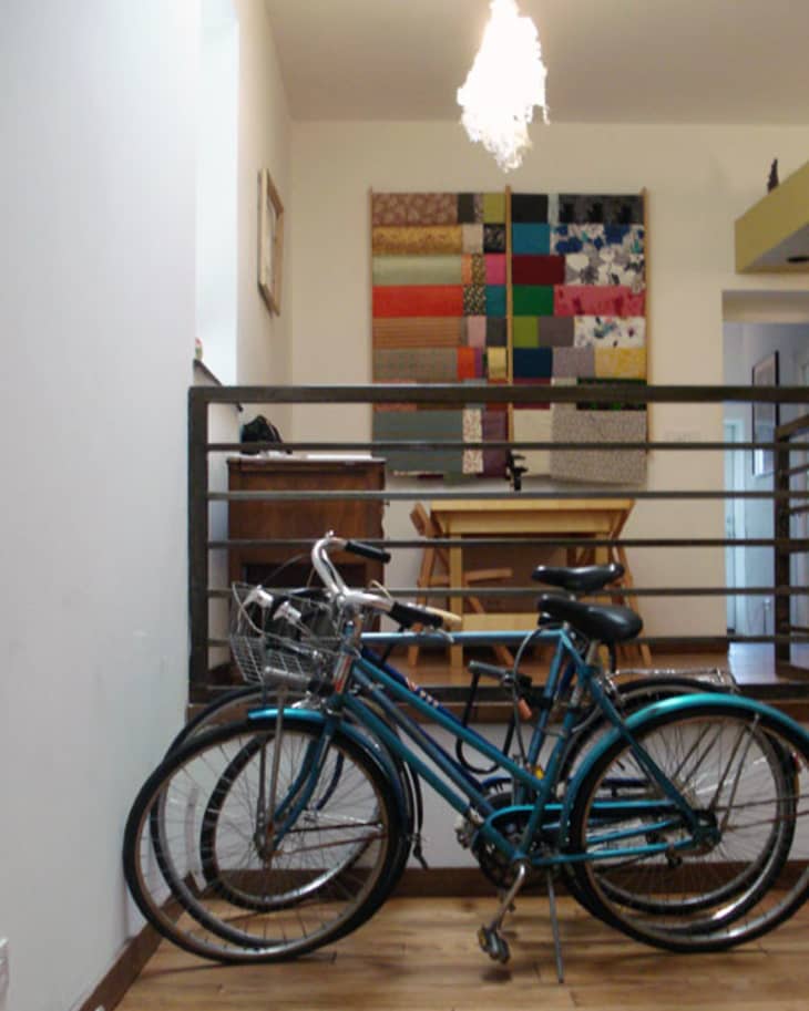 Two blue bicycles parked indoors near a wooden desk and chair, with a colorful patchwork quilt on the wall.