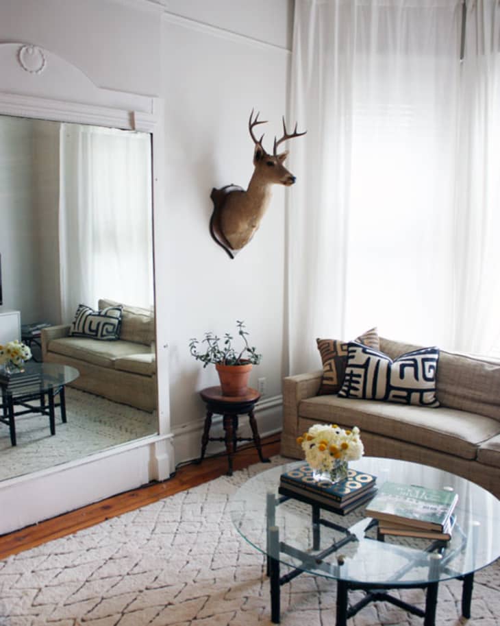 Living room with beige sofa, geometric pillows, glass coffee table, deer head mount, and large mirror reflecting decor.