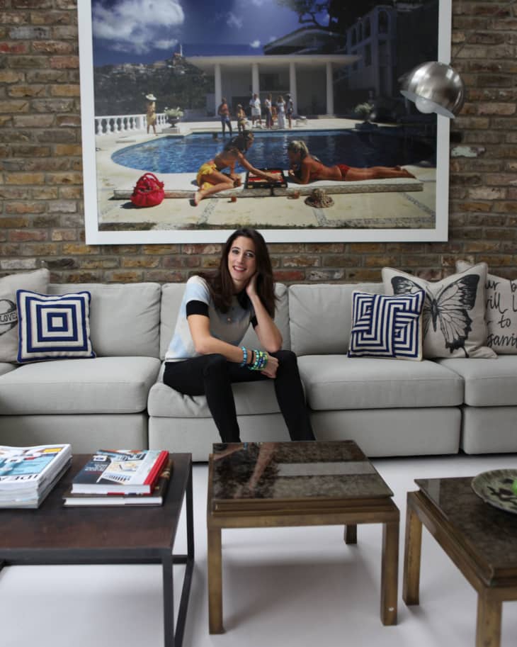 Woman sitting on a gray sofa with geometric pillows, brick wall, and large poolside photo above.