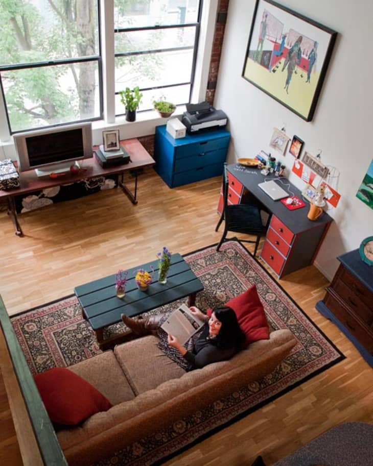 Loft-style living room with large windows, a beige sofa, colorful cushions, a patterned rug, and a desk with a laptop.