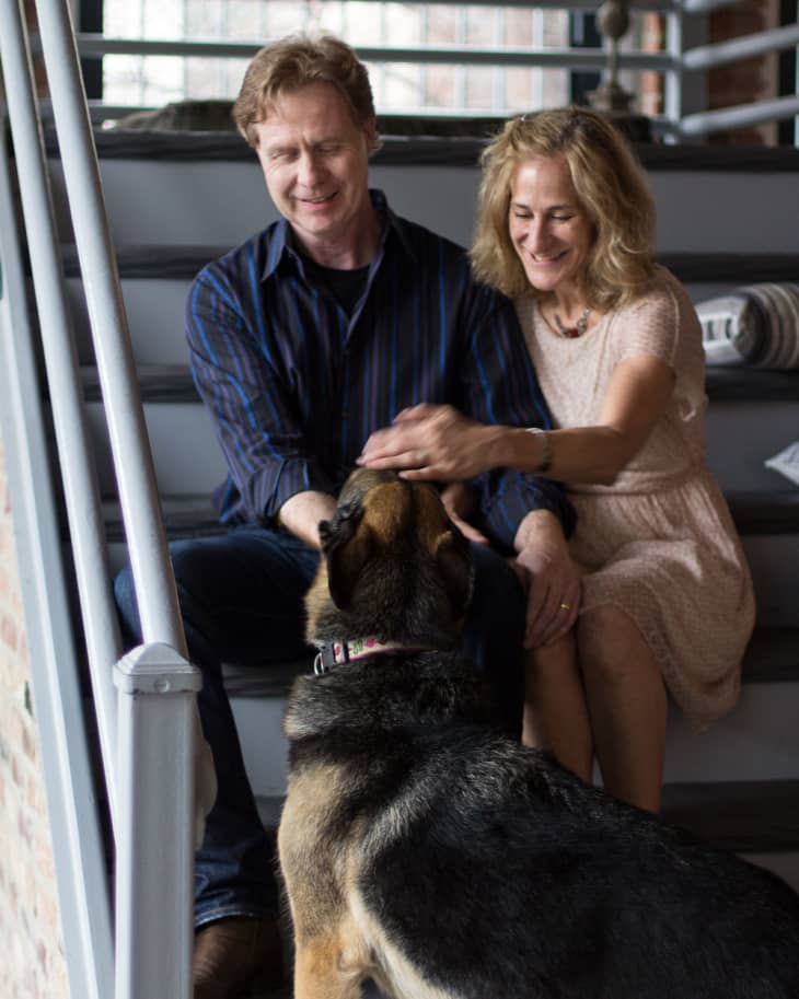 Couple sitting on stairs petting a German Shepherd dog indoors.