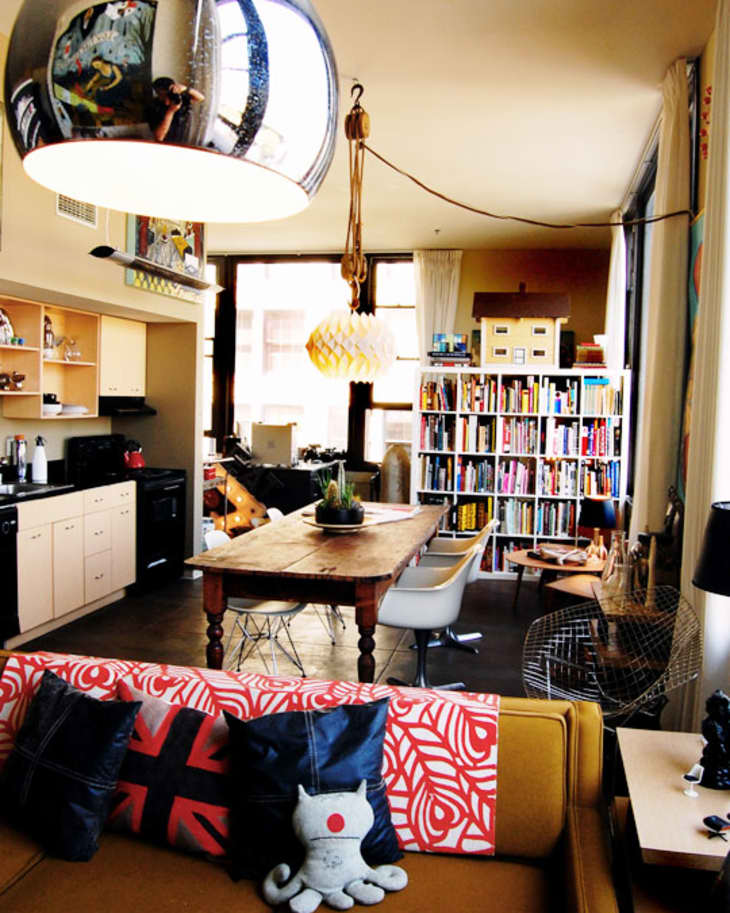Loft apartment with a mustard sofa, colorful pillows, wooden dining table, bookshelves, and modern pendant lights.
