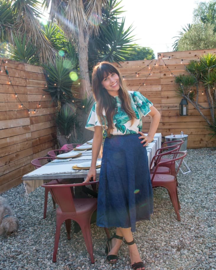 Woman in a tropical print top and blue skirt standing by an outdoor dining table with red chairs and string lights.