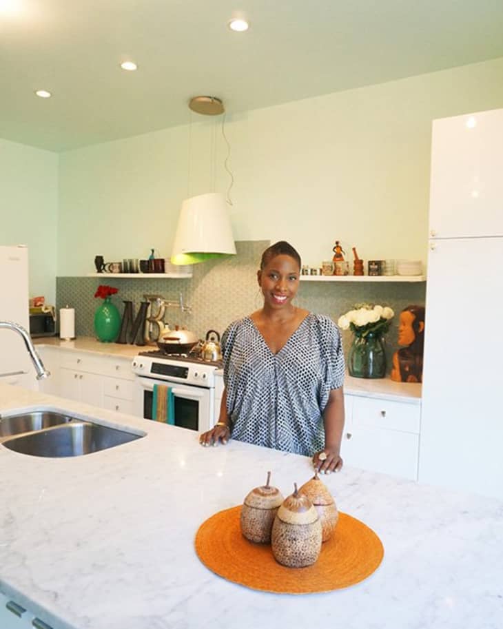 Woman standing in a modern kitchen with white cabinets, marble countertop, and decorative items.