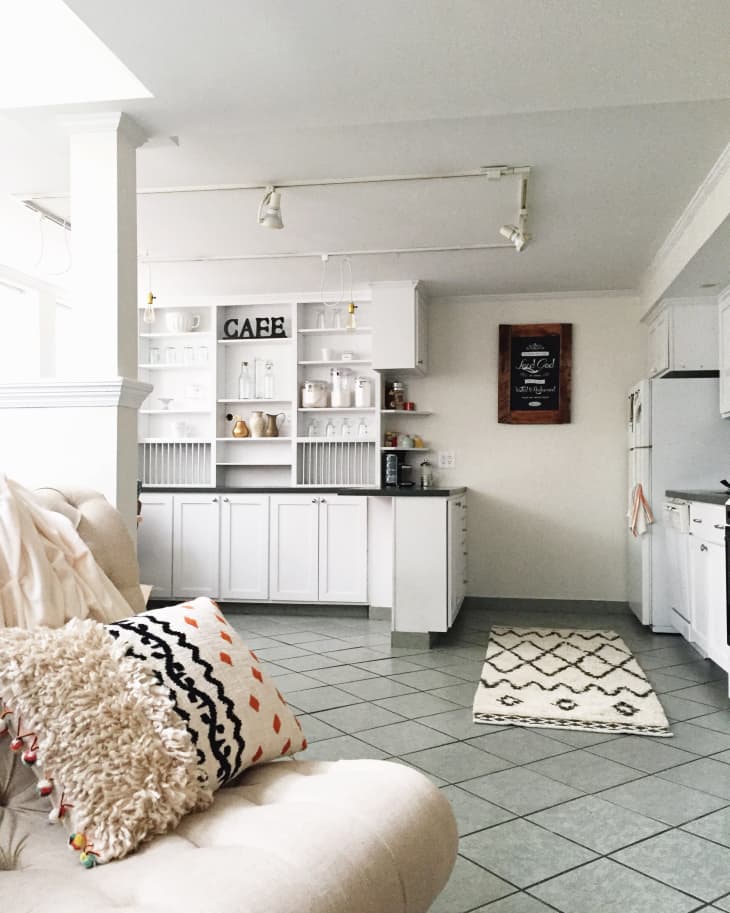 Open kitchen with white cabinets, a "CAFE" sign, and a chalkboard. Cream sofa with patterned pillows in the foreground.