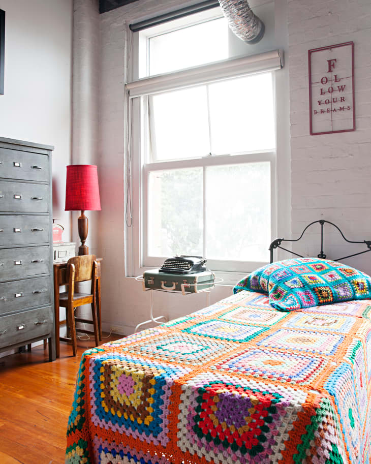 Bedroom with colorful crochet bedspread, vintage typewriter on suitcase, red lamp, and "Follow Your Dreams" sign.